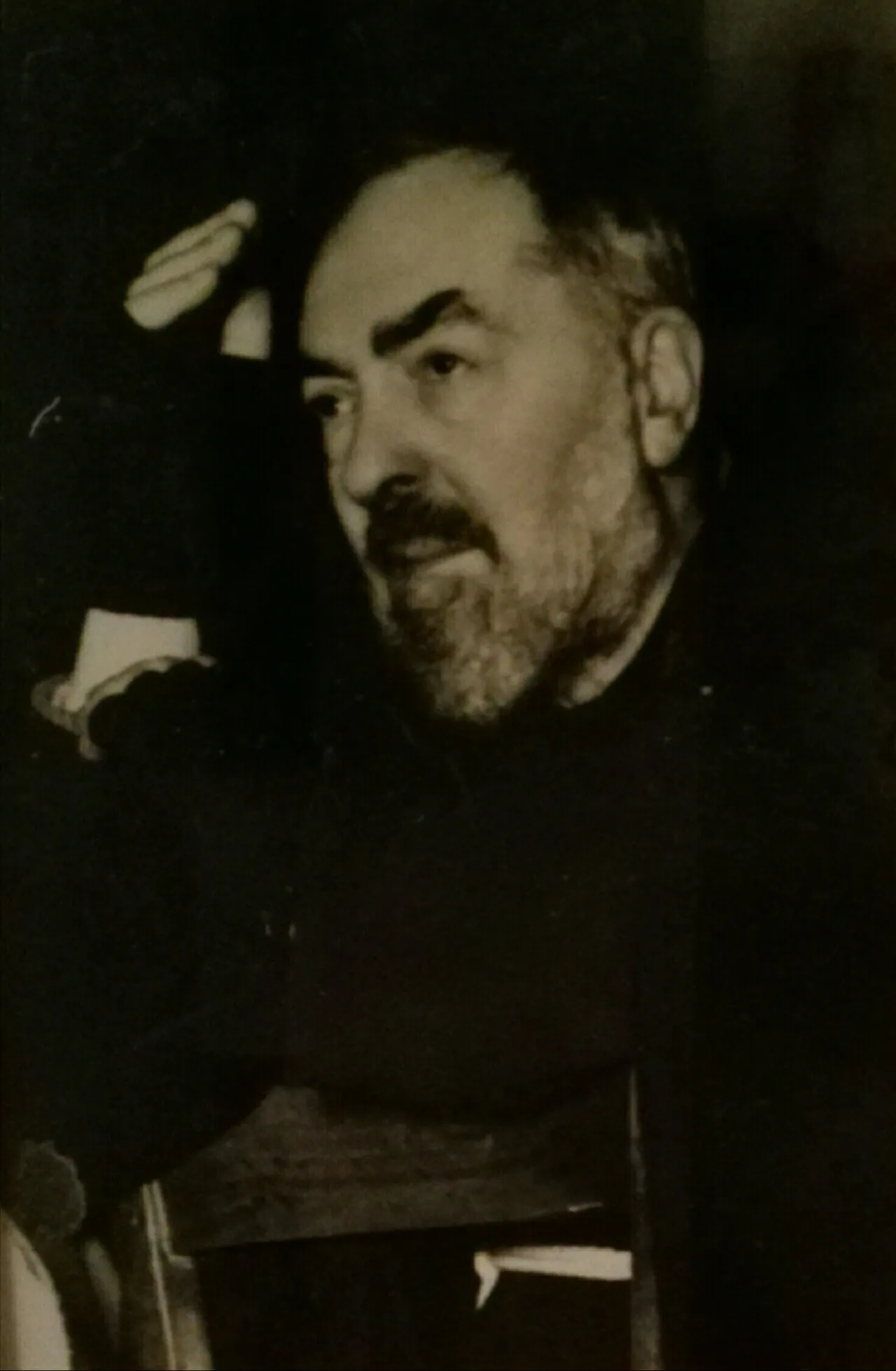 Fotografia de Padre Pio com hábito capuchinho, sentado e apoiando uma das mãos na cabeça.